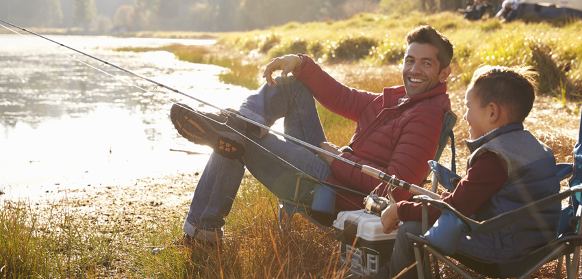 An adult man and a young boy sit in folding chairs on a grassy bank, smiling while fishing together at the edge of a calm lake during a sunny afternoon - https://www.loyaltyinsurance.com/