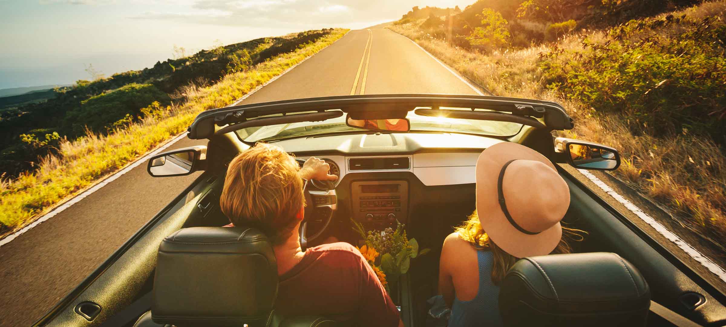 Rear view of a couple driving a convertible down a long, scenic road toward a golden sunset - https://www.loyaltyinsurance.com/